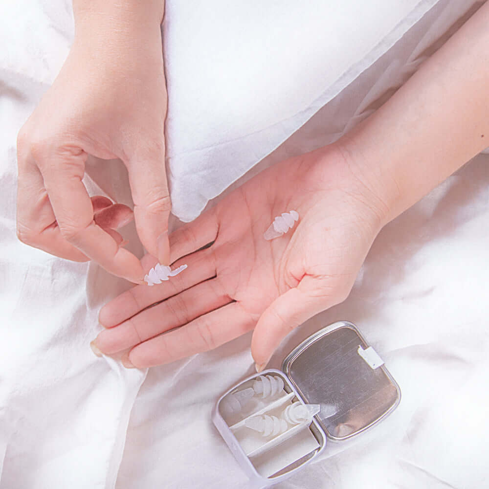 Kokon Universe noise reducing earplugs held in hand, with a soft case on a pillow background for peaceful sleep.