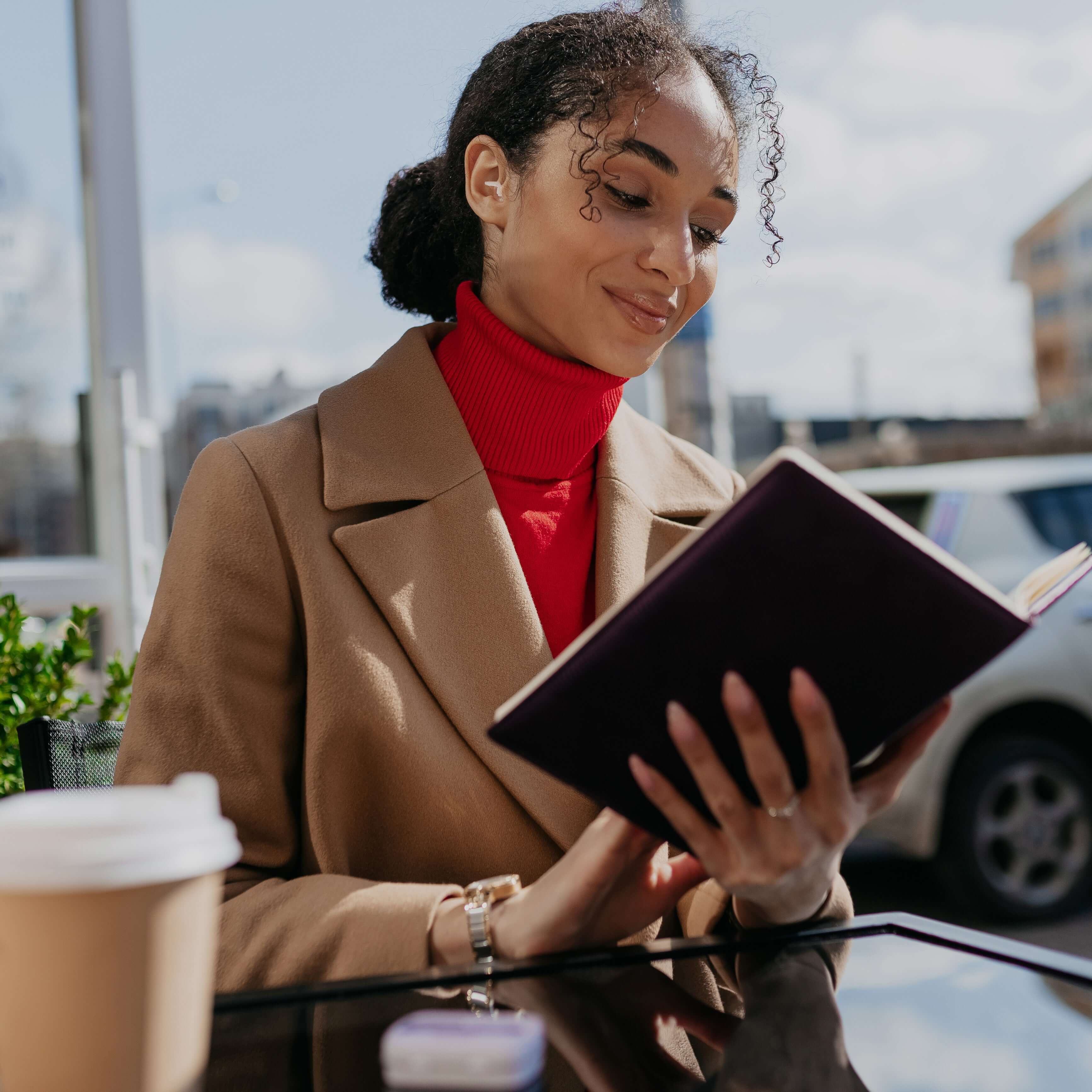 Woman in a stylish coat reading a book at an outdoor café with a coffee cup.