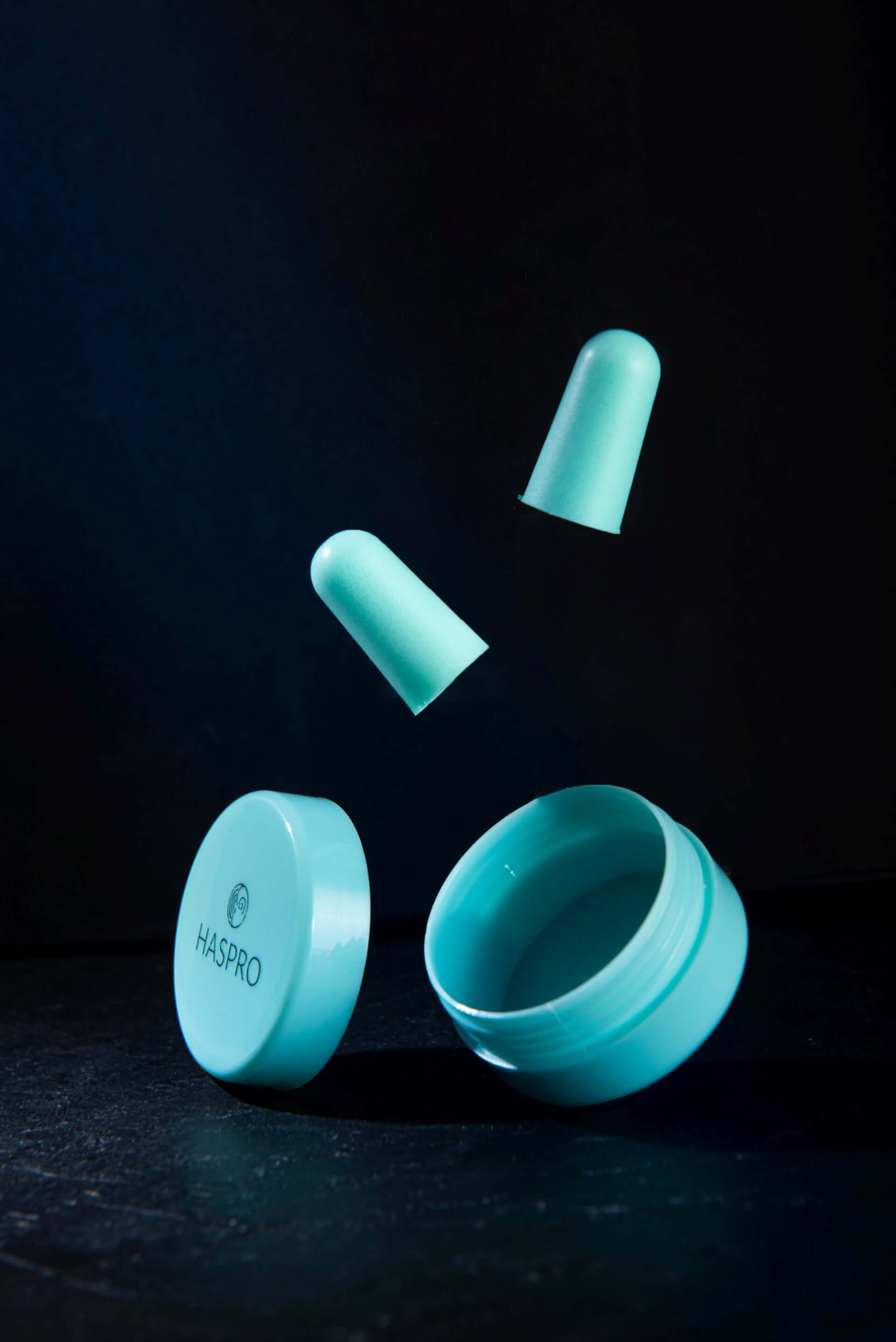 Turquoise HASPRO earplug case with two foam earplugs floating above it, designed for high noise reduction.