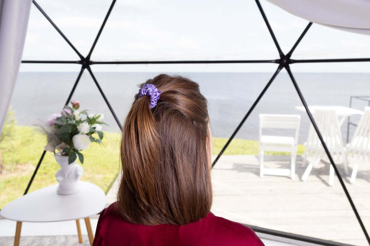 Woman with brown hair styled in a ponytail using a purple Pure Mulberry Silk Scrunchie by a large window overlooking the water.
