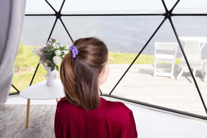 Woman with hair in a ponytail wearing a purple pure mulberry silk scrunchie, seated by a window with a scenic view.