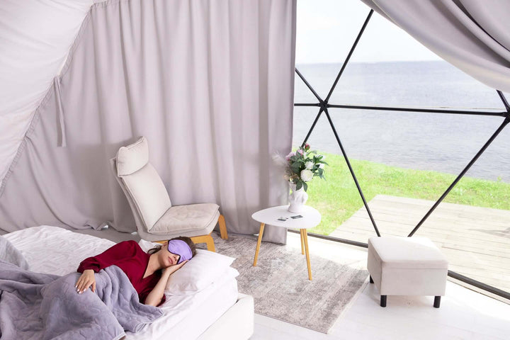 Woman sleeping peacefully with Quietstore 3.0 silk sleep mask in a serene bedroom setting by the water.