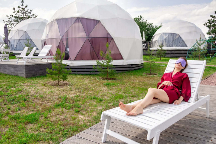 Person relaxing outdoors in a silk sleep mask near geodesic domes, embodying comfort and tranquility.