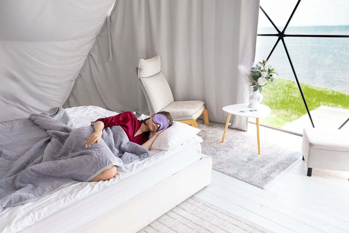 Person wearing a silk sleep mask relaxing on a bed in a modern, serene bedroom setting with natural light.