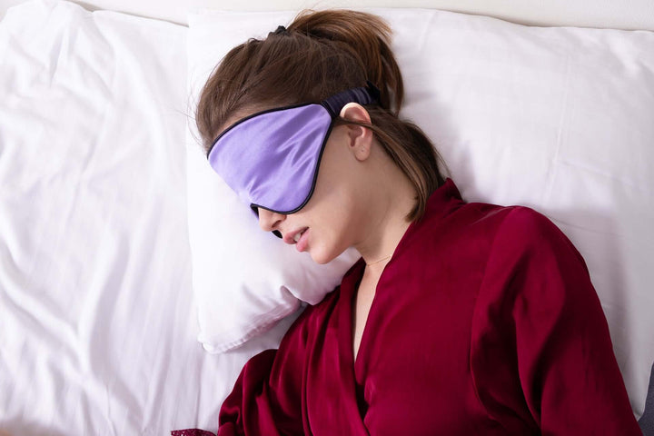 Woman sleeping with Quietstore 3.0 silk sleep mask on a pillow, wearing a red robe for a restful night's sleep.
