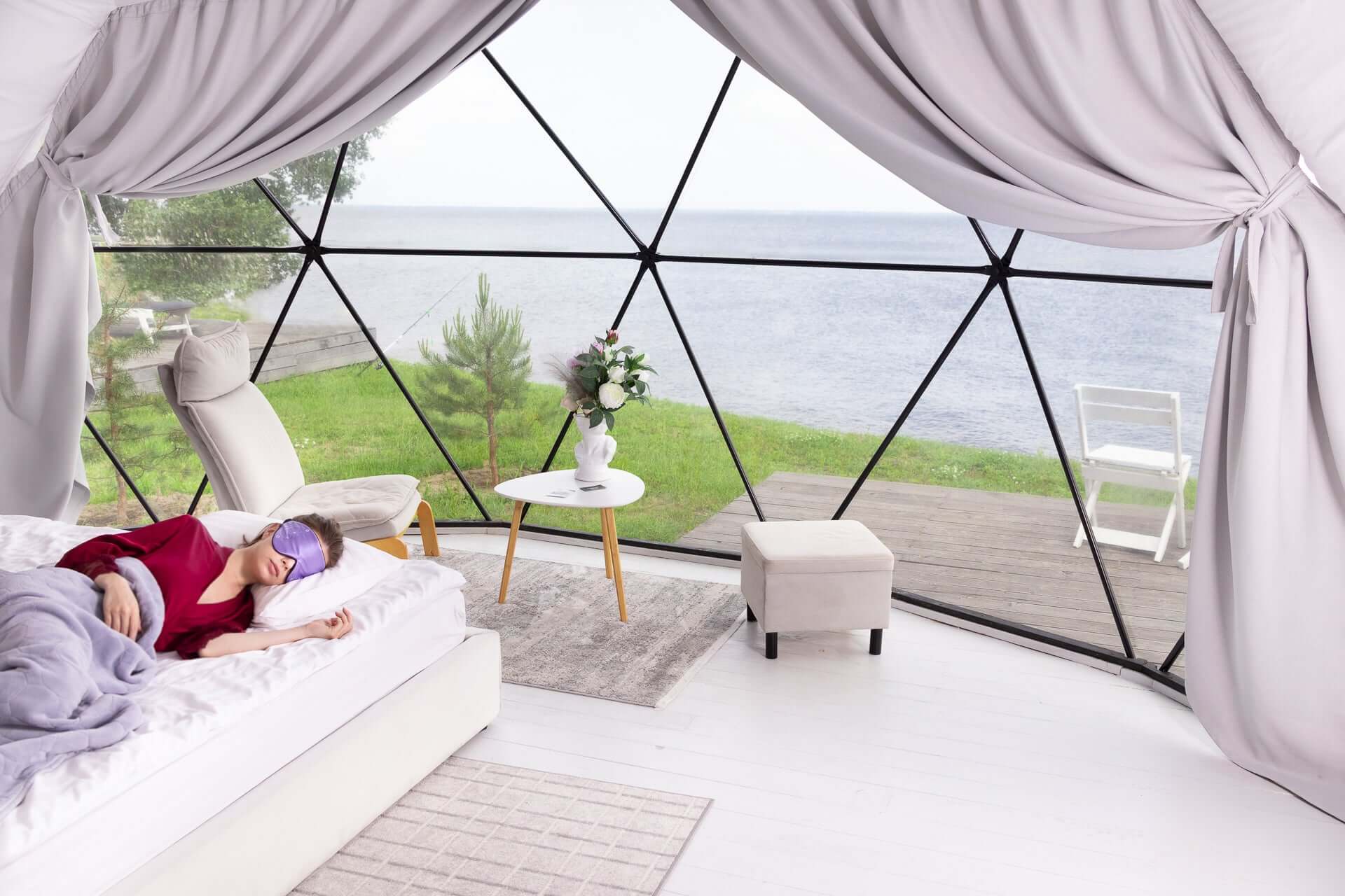 Woman relaxing in a cozy bedroom with Quietstore silk sleep mask, overlooking a serene lakeside view.
