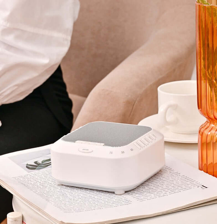 White Noise Sound Machine on a coffee table next to a person, perfect for masking distracting sounds at home.