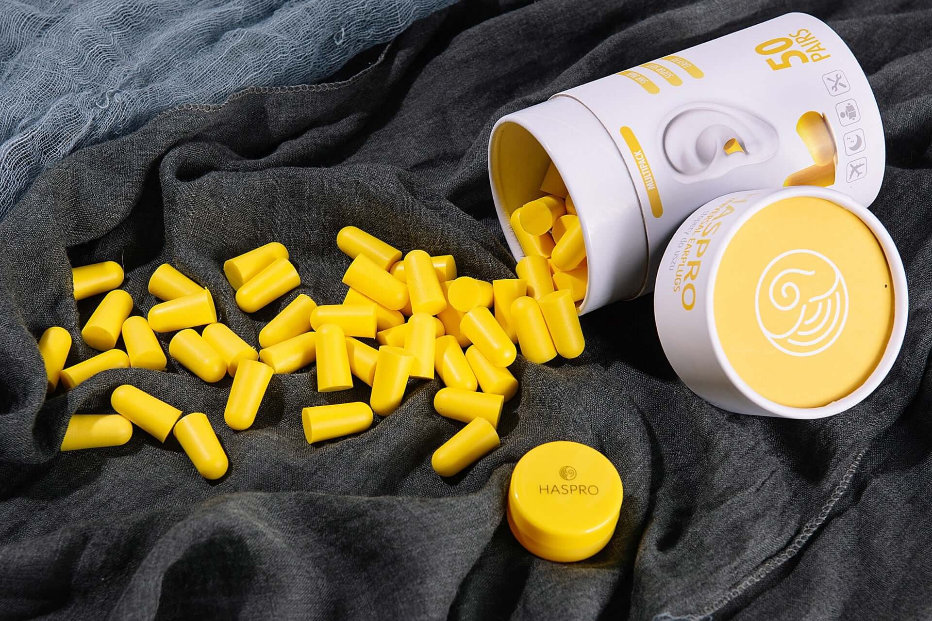 HASPRO Tube50 foam earplugs in yellow spilling from a storage container on a dark fabric background.